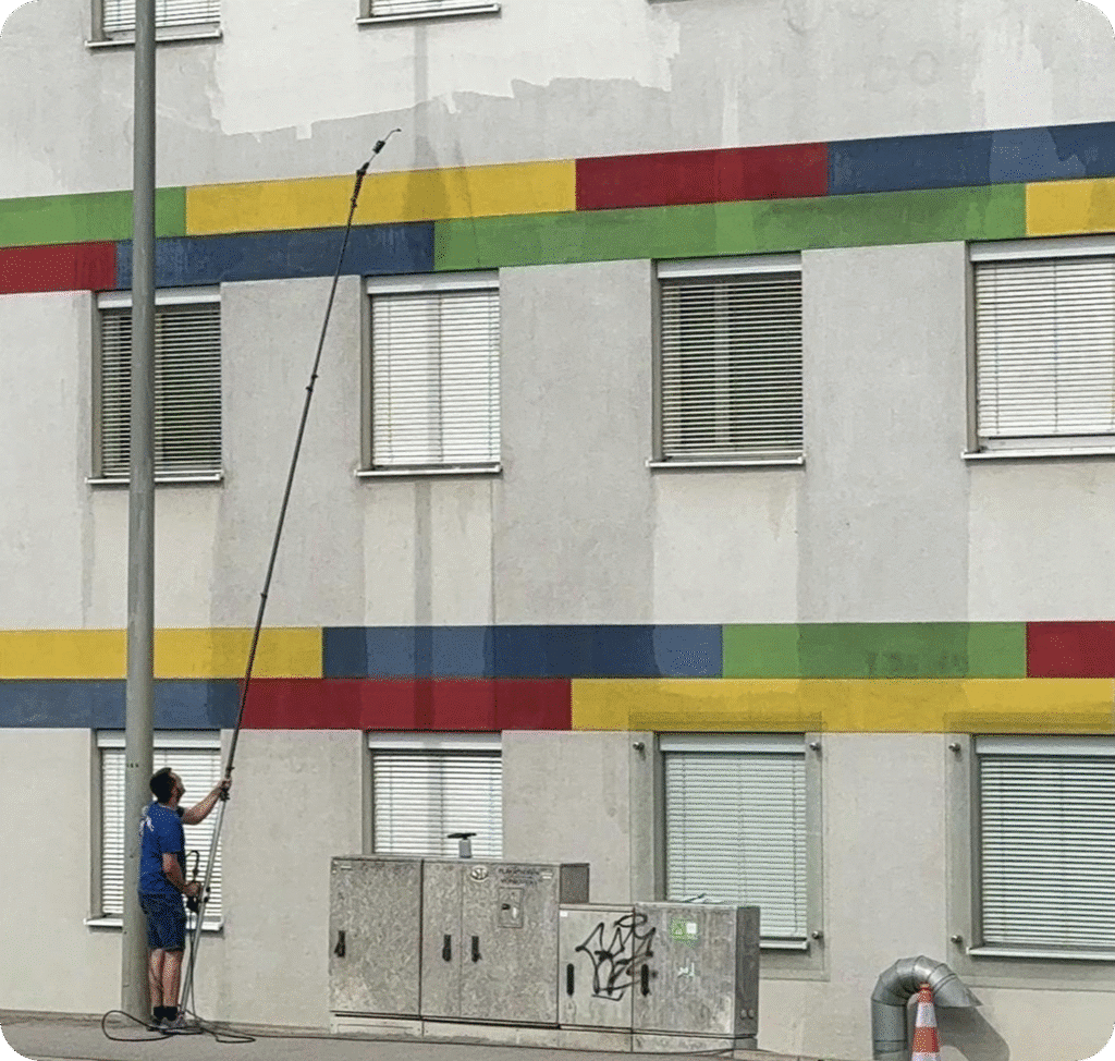 Reinigungskraft säubert mehrfarbige Gebäudefassade mit langem Teleskopstab – QClean Fassadenreinigung